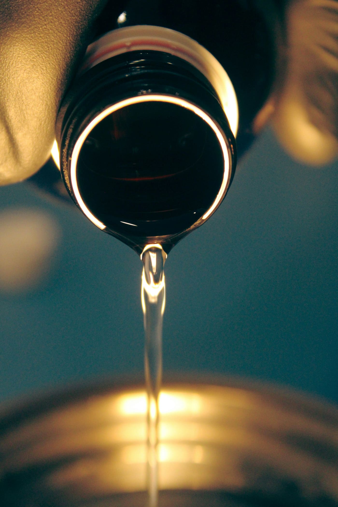 Hilo de aceite dorado cayendo a contraluz desde el cuello de una botella oscura.
