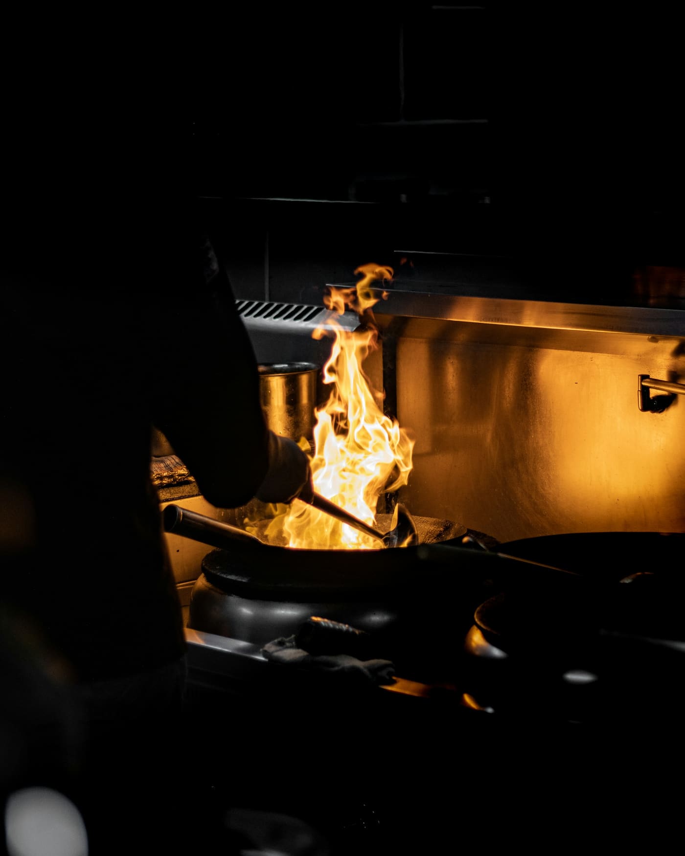 Cocina profesional casi a oscuras: una llamarada dorada se levanta desde un wok mientras se saltea.